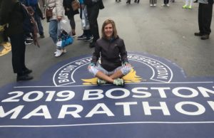 A woman sitting on the ground in front of a boston marathon sign.