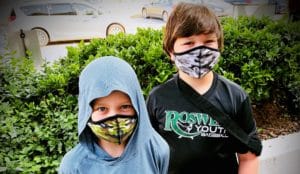 Two boys wearing masks standing next to a bush.